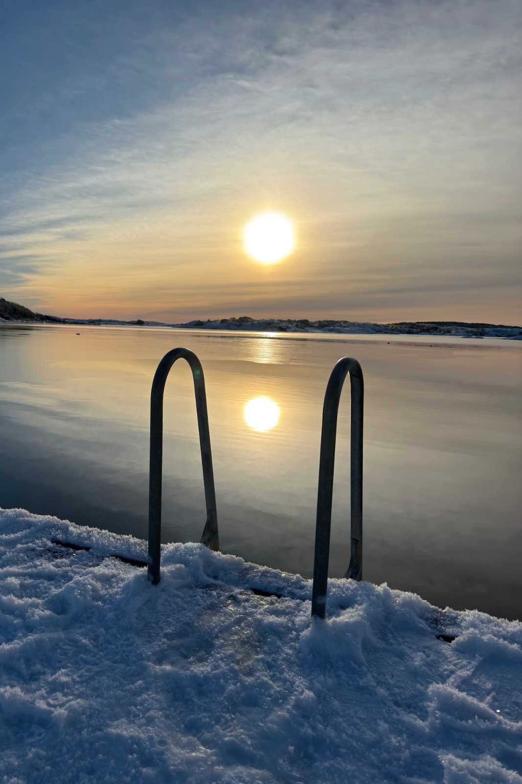 Badbrygga med snö och stege där solen speglar sig i vattnet